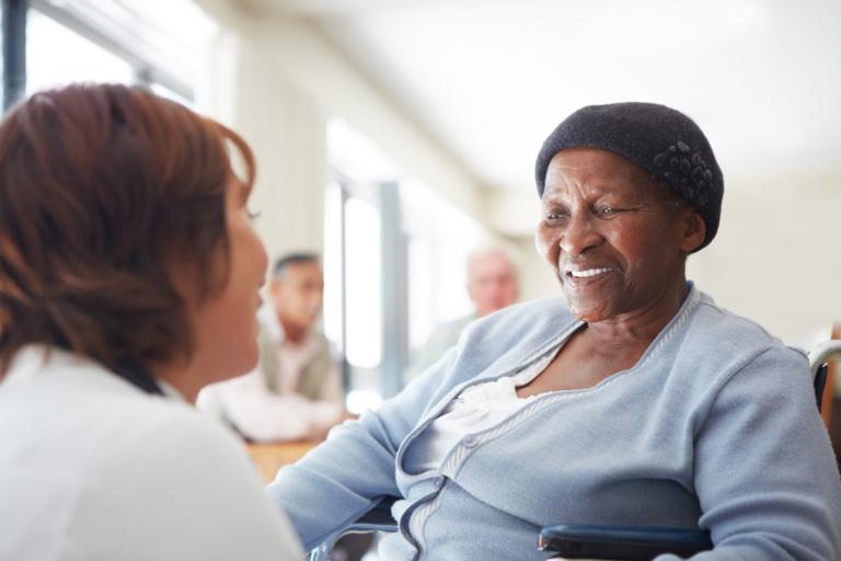 A woman wearing a black hat and light blue cardigan is smiling at another person who is partially visible and facing her. The background is softly blurred, featuring other people and a bright, naturally lit room.