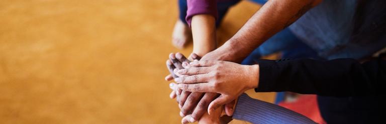 A group of hands of different skin tones are stacked on top of each other in a show of unity and teamwork against a blurred, warm-colored background.